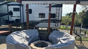 Un espace extérieur aménagé avec des sièges, protégé par un auvent, comprenant un foyer, entouré de tables et de chaises également protégées par un auvent, le tout sous un pavillon d’où l’on peut apercevoir un camping-car au loin.