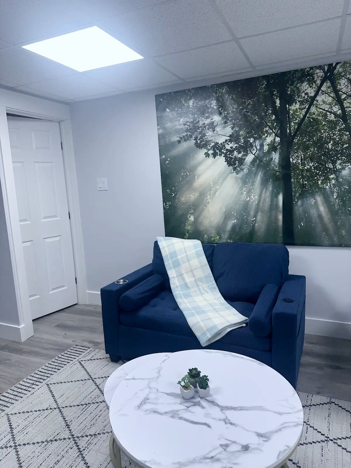 Una habitación con un sofá azul, una mesa redonda blanca con encimera de mármol y un mural en la pared que representa un bosque iluminado por el sol.