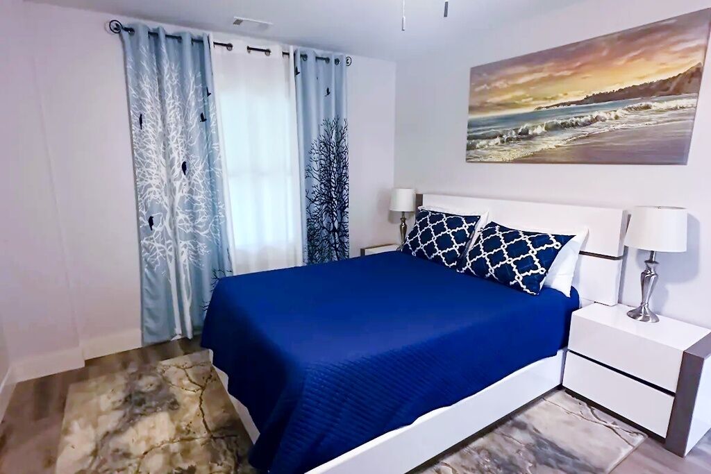 Un dormitorio con cama, cortinas, mesita de noche, lámpara y un cuadro en la pared.