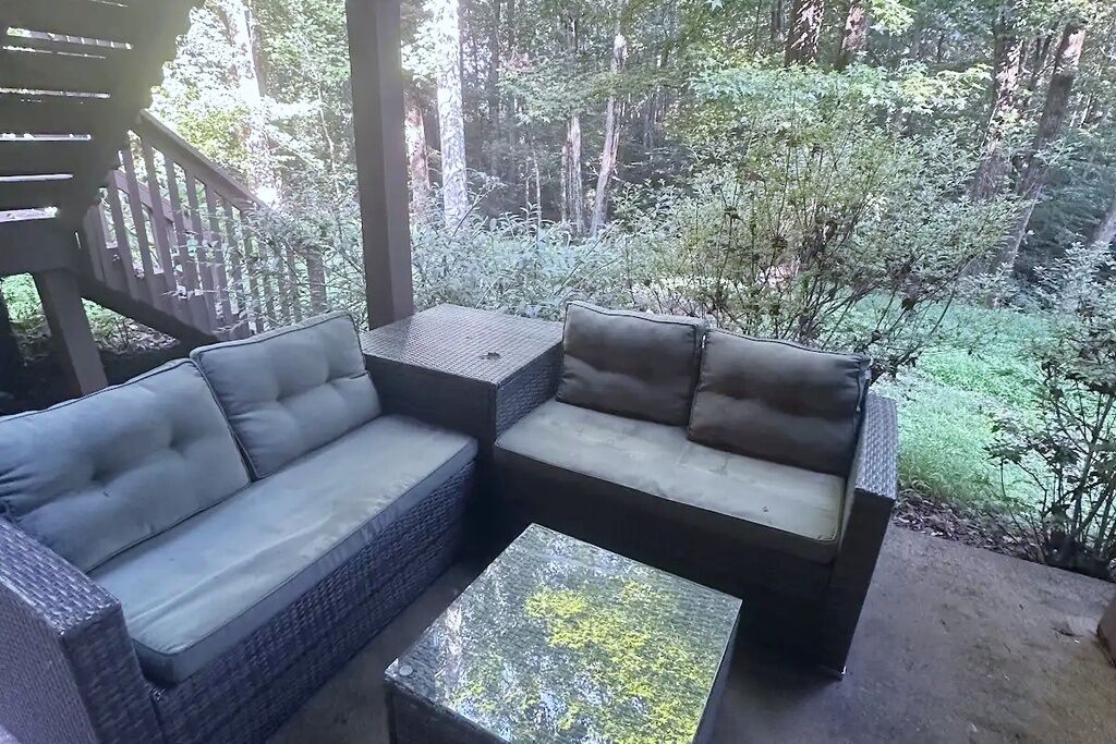 Un área de estar al aire libre con sofá y una mesita, rodeada por una terraza de madera y un paisaje boscoso.