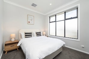 Un dormitorio con un ventanal grande, una cama con ropa de cama blanca, dos mesitas de noche con lámparas y una obra de arte colgada en la pared.
