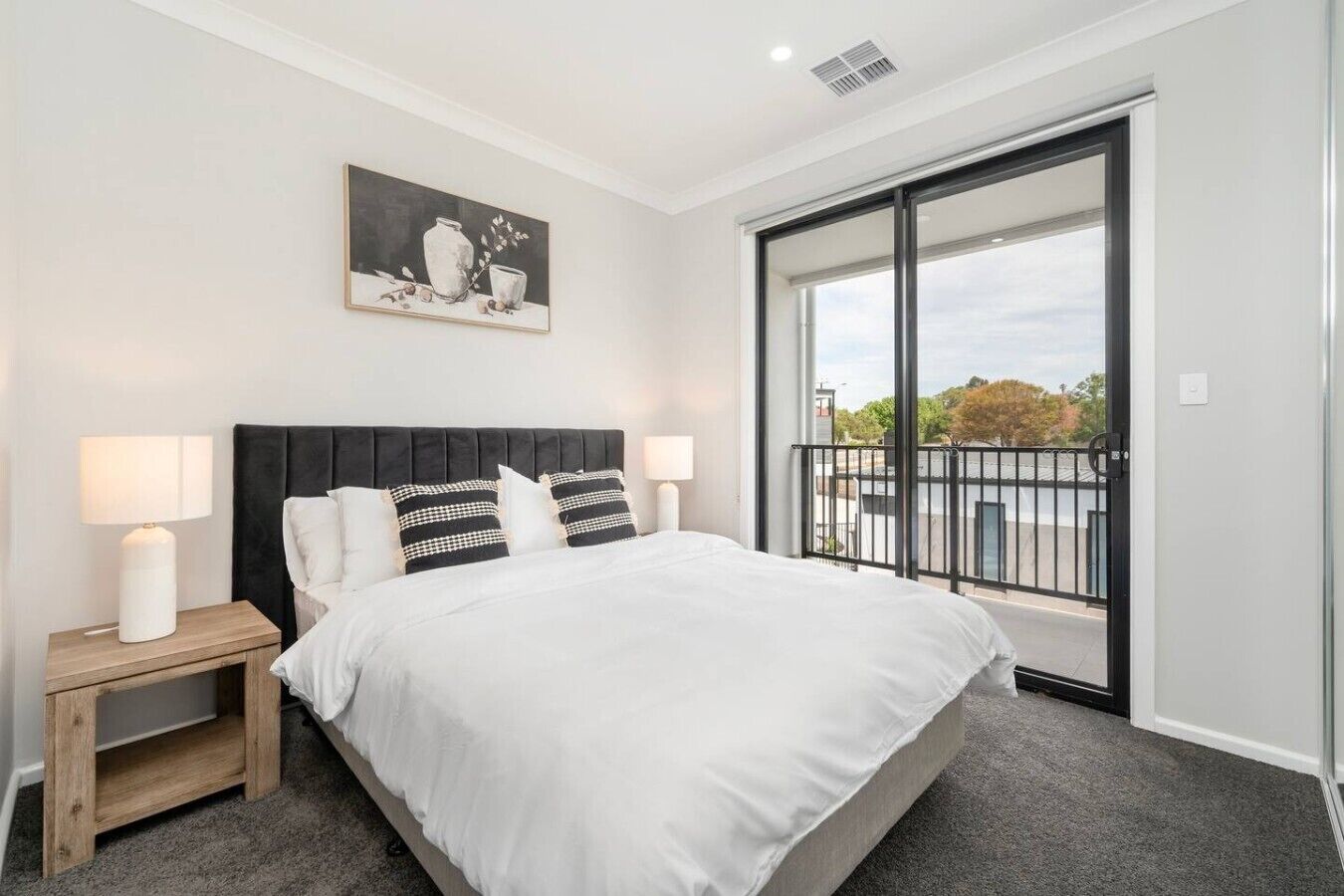 A bedroom with a large bed, two bedside tables with lamps, a black headboard, a painting on the wall, and a balcony with a view.