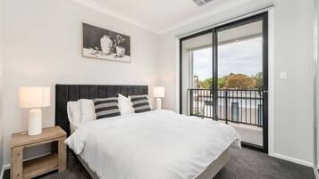 A bedroom with a large bed, two bedside tables with lamps, a black headboard, a painting on the wall, and a balcony with a view.