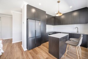 Una cocina moderna con armarios oscuros, un refrigerador de acero inoxidable, una isla central con un taburete y una lámpara colgante.