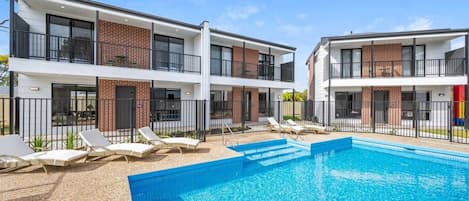 Um edifício moderno com piscina, espreguiçadeiras e área cercada.