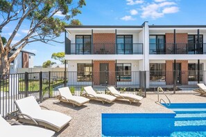A two-story building with a swimming pool, lounge chairs, and a fenced garden area.