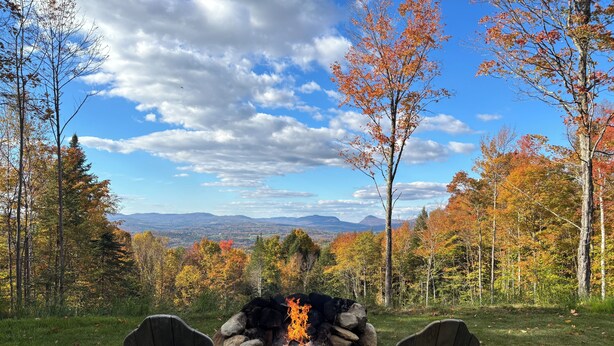Un área boscosa con un hogar para fuego y dos sillas Adirondack mirando un paisaje con montañas y árboles.