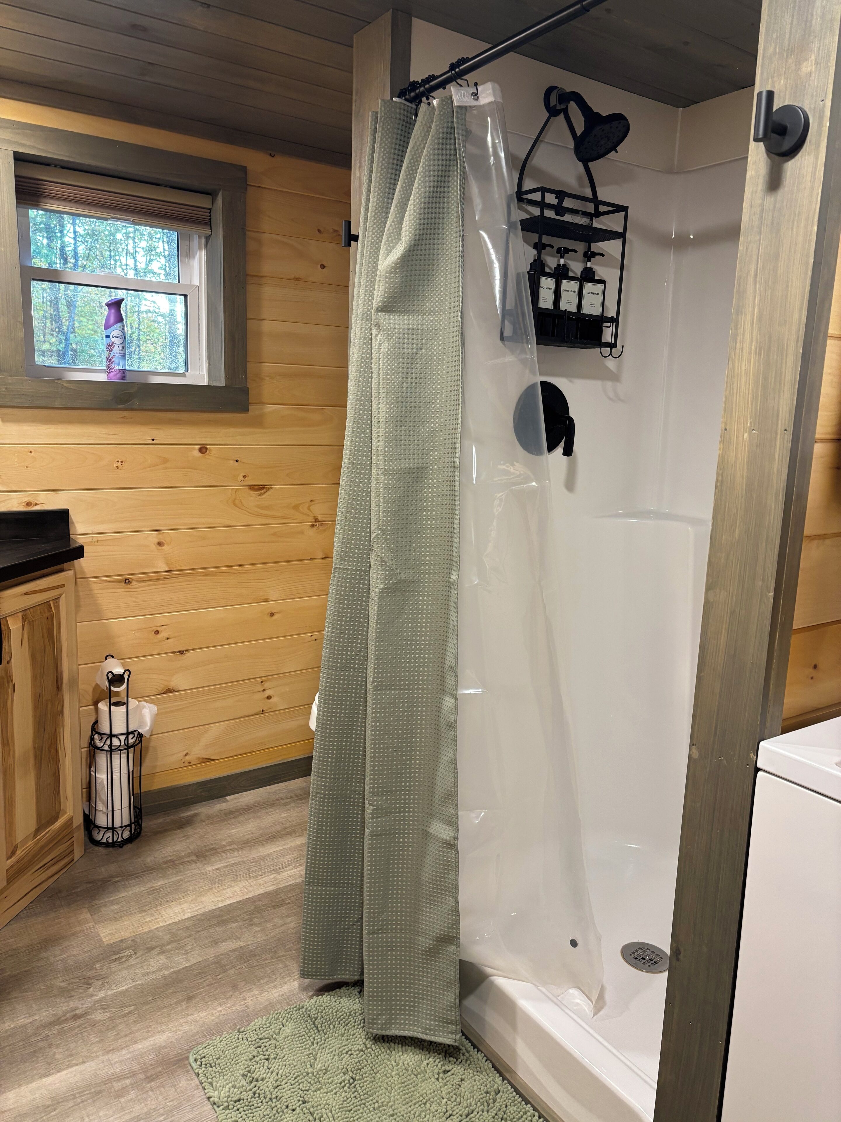 Suihkussa on läpinäkyvä verho, seinät ovat puuta, ikkunasta avautuu näkymä puihin, ja siellä on pieni hylly, jossa on hygieniatuotteita.