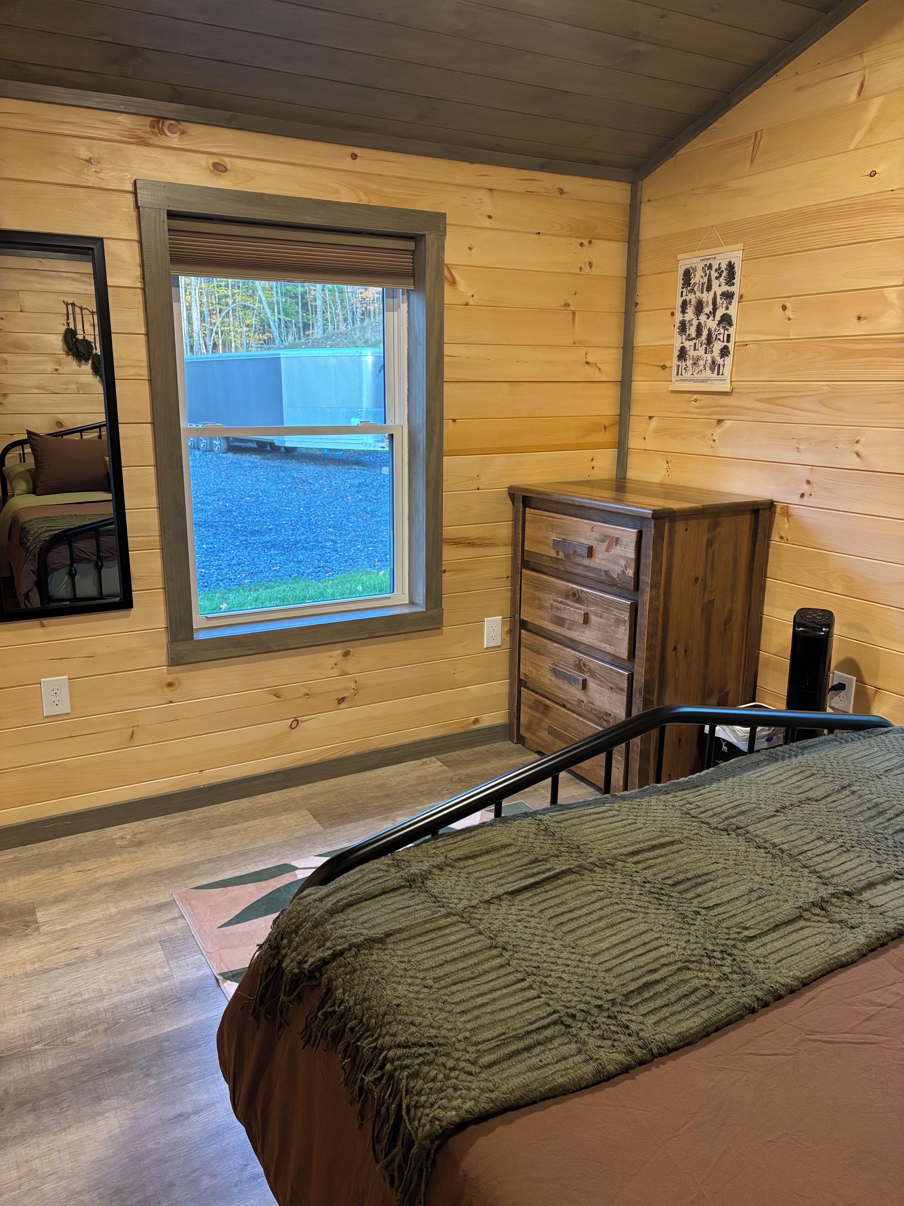 A wooden cabin room with a bed, dresser, and a window overlooking a gravel path.