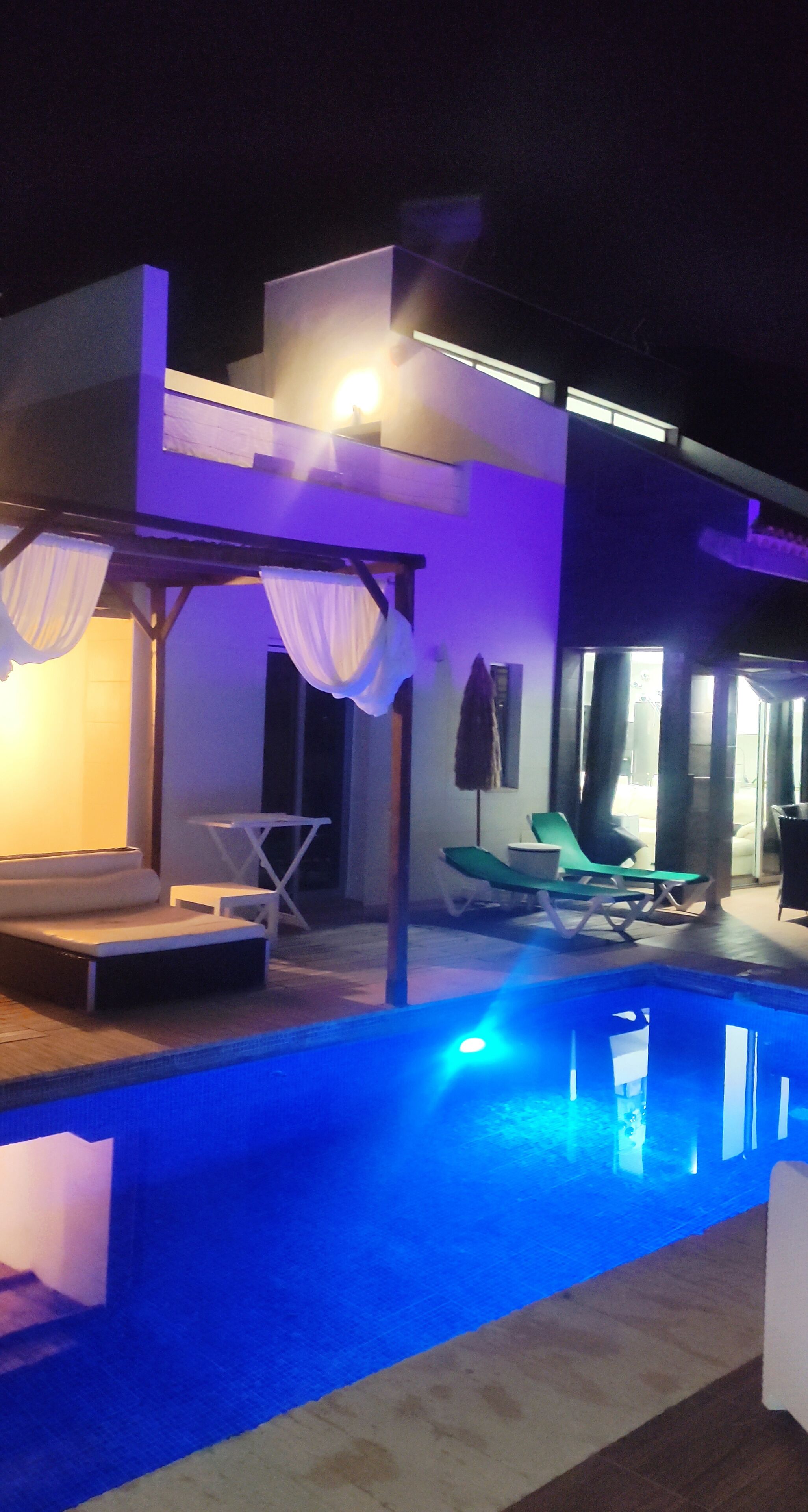 Uma área de piscina moderna com piscina iluminada em azul, espaço de estar coberto com cortinas brancas e uma espreguiçadeira.