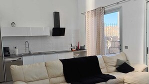Un salon moderne avec un canapé beige, une table basse blanche et un coin cuisine doté de meubles blancs et d’une hotte noire.