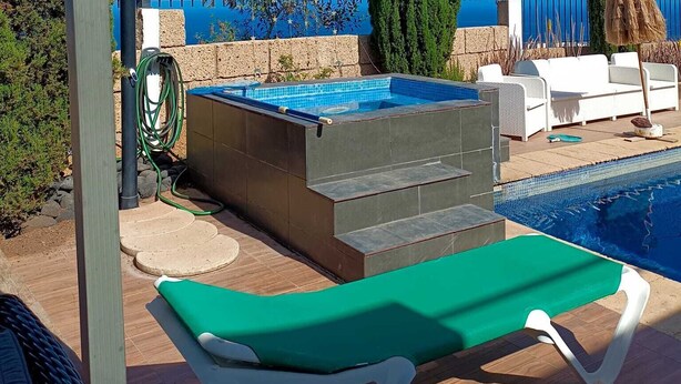 Une piscine avec un fauteuil de jardin vert, un tuyau d’arrosage et de grands cyprès en arrière-plan.