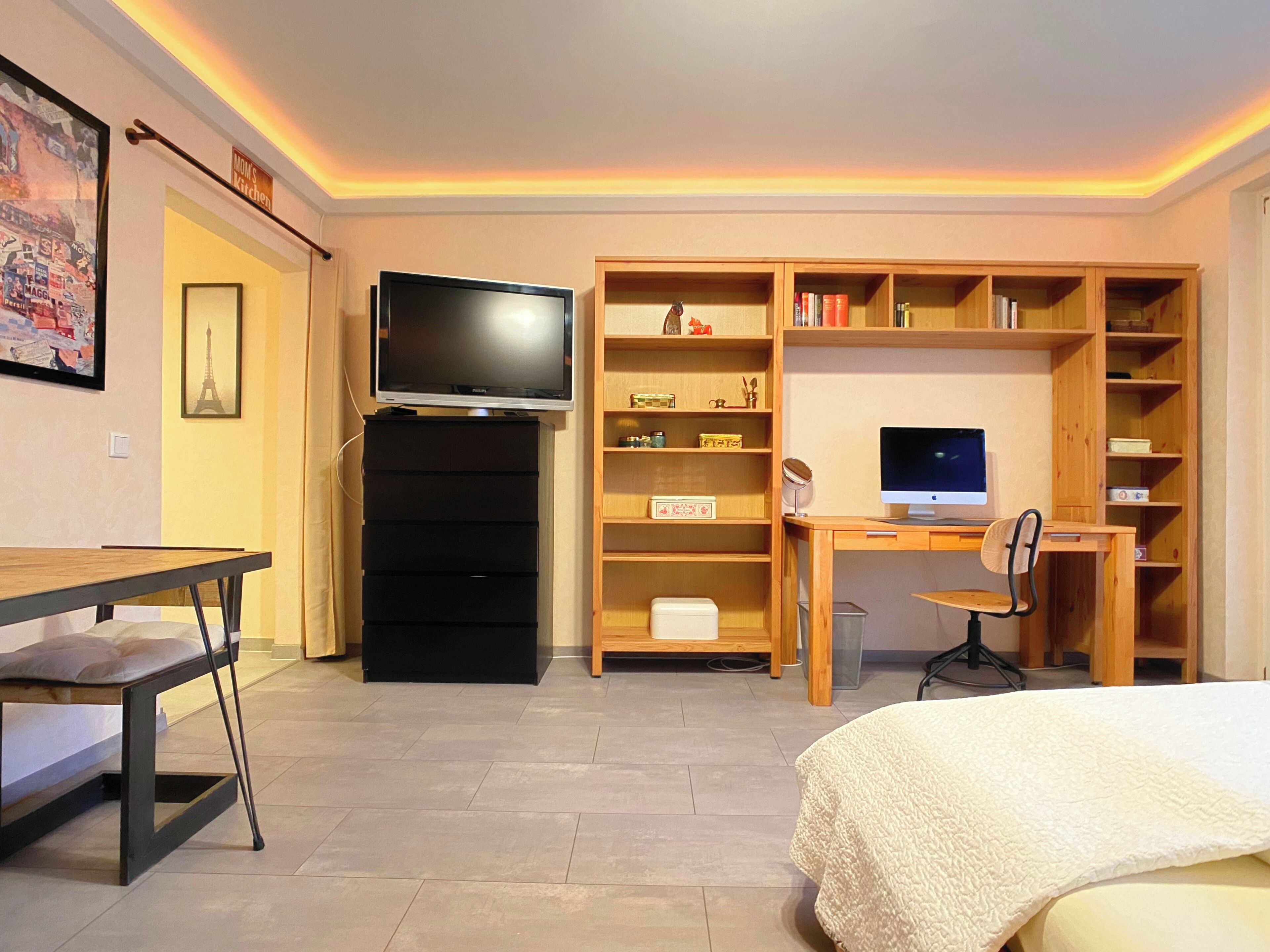 A hotel room with a bed, a desk with a computer, a TV, a bookshelf, and a small table.