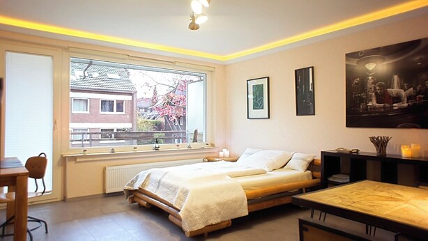 Une chambre avec un lit, une fenêtre donnant sur une maison et des cerisiers en fleurs, une table et une chaise.