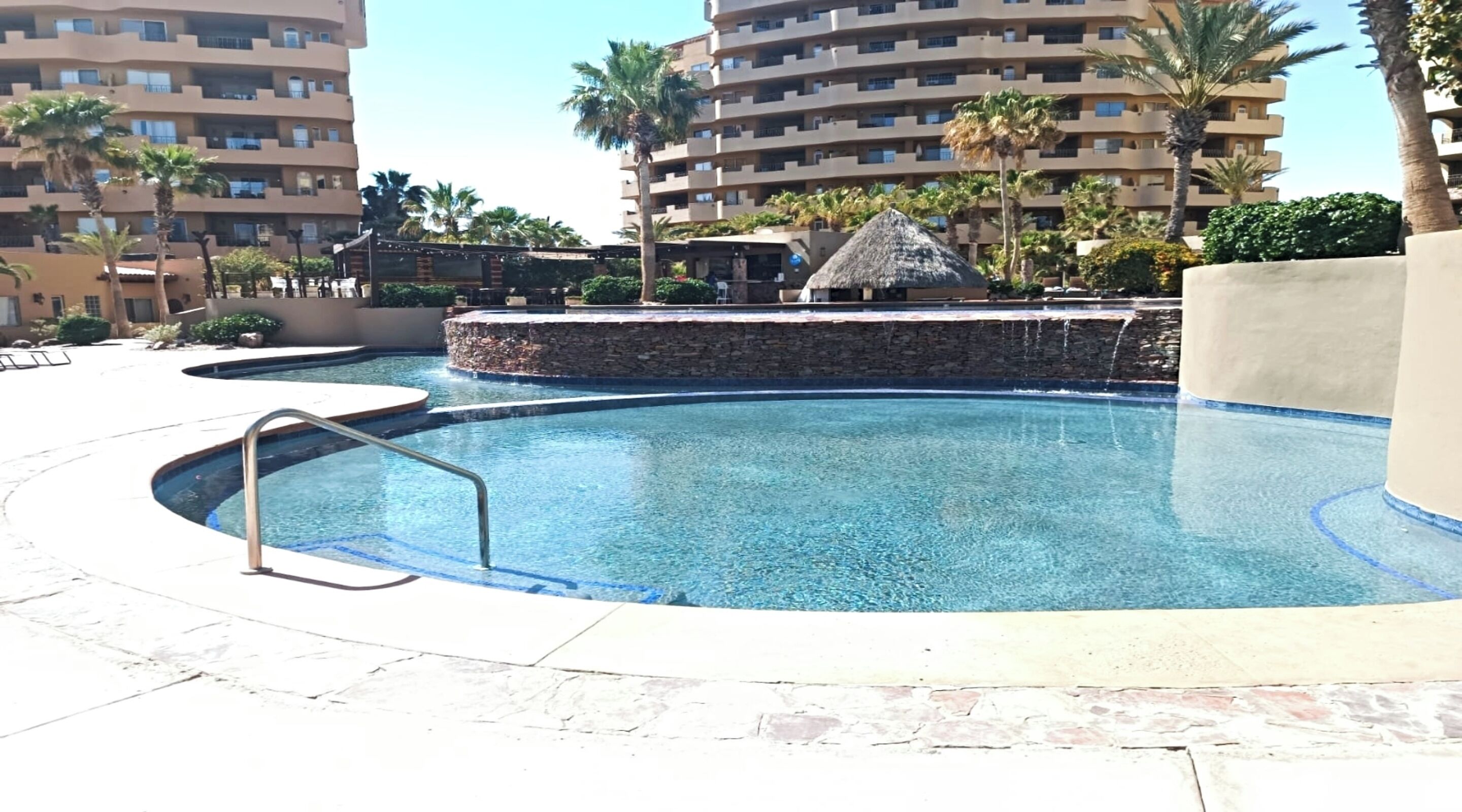 A swimming pool with a stone wall and a thatched-roof structure, surrounded by palm trees and a multi-story building with balconies.