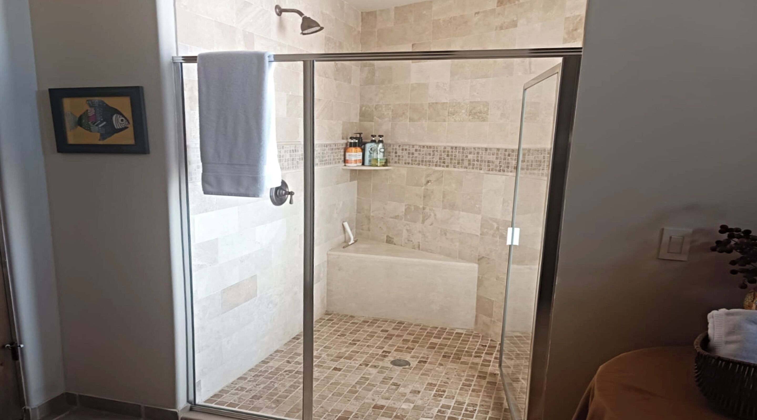 Zona de ducha con paredes de vidrio, bañera, paredes con azulejos y un estante con artículos de aseo.