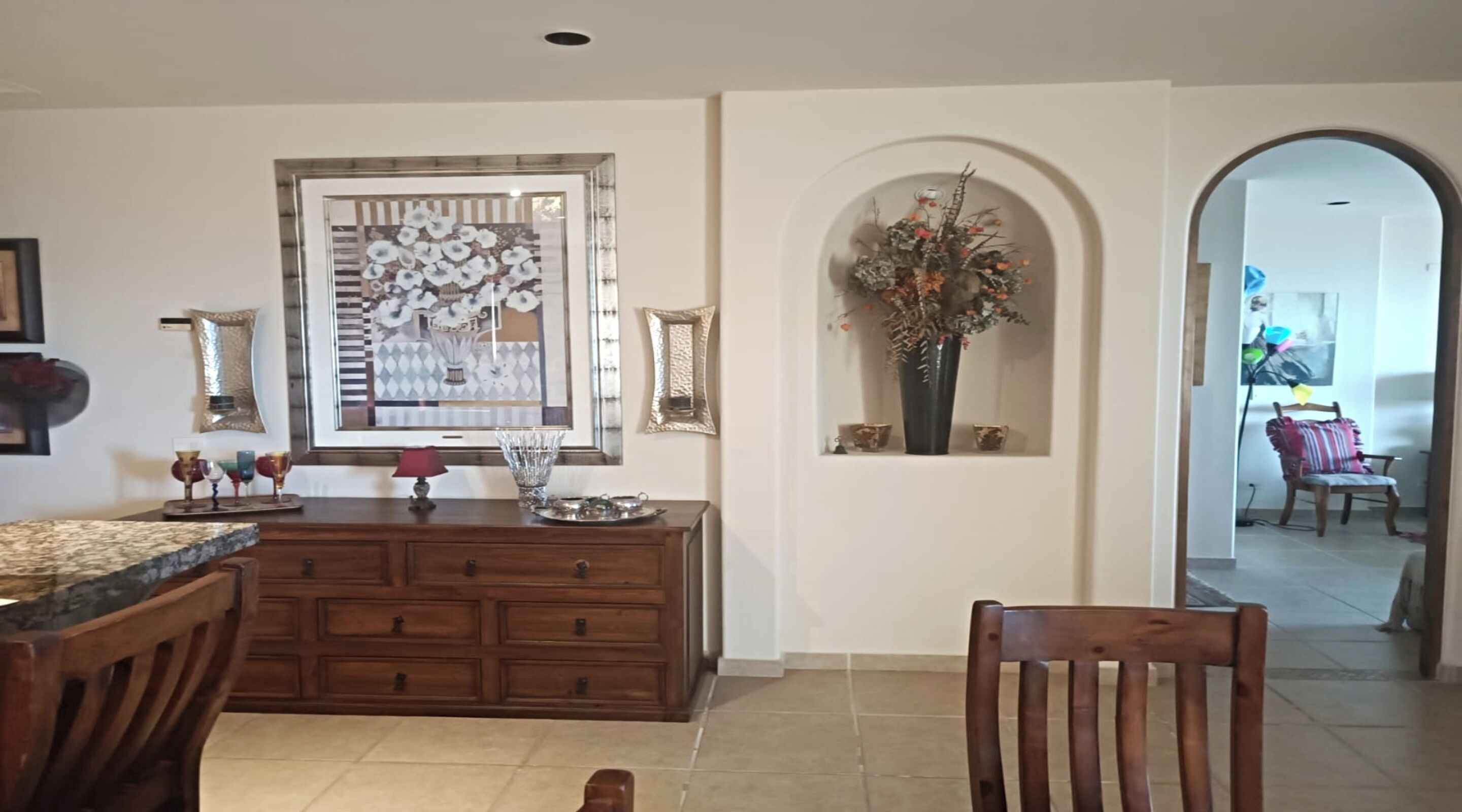Un coin repas avec une table et des chaises en bois, un miroir décoratif et un vase avec des fleurs accroché au mur.