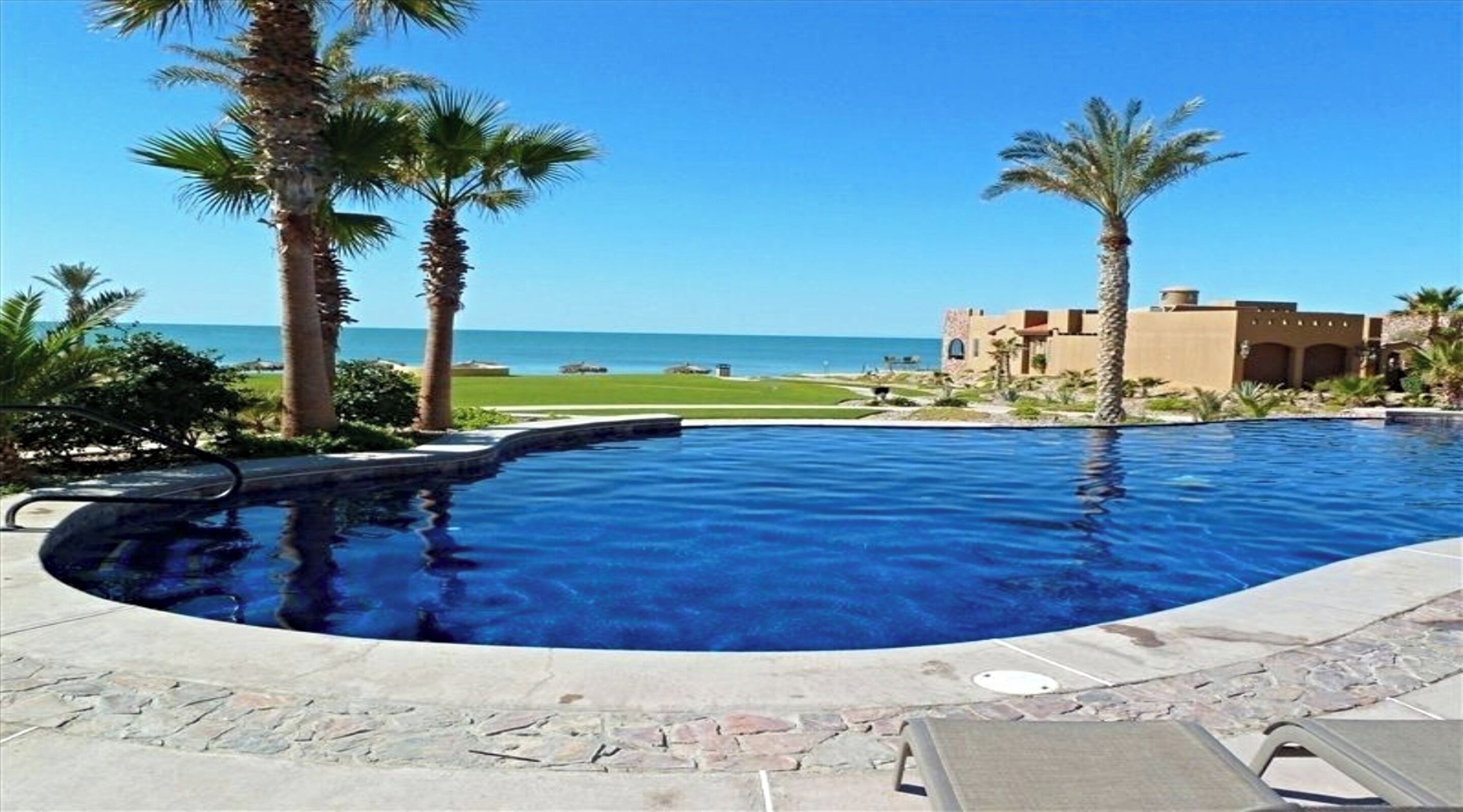 A swimming pool with a view of the ocean, surrounded by palm trees and a building with a desert-style architecture.