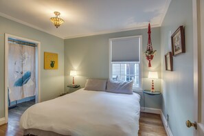 A bedroom with a large bed, two bedside tables with lamps, a window with blinds, a door with a curtain, and a wall with a framed picture. - East Nashville Bamboo Cottage (Nashville)