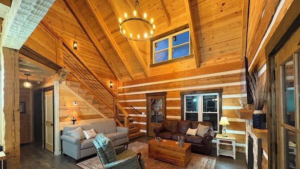 A log cabin interior with a staircase, two sofas, a wooden coffee table, and a chandelier.