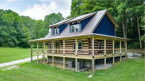 A two-story log cabin with a wooden deck and a metal roof, surrounded by lush greenery.