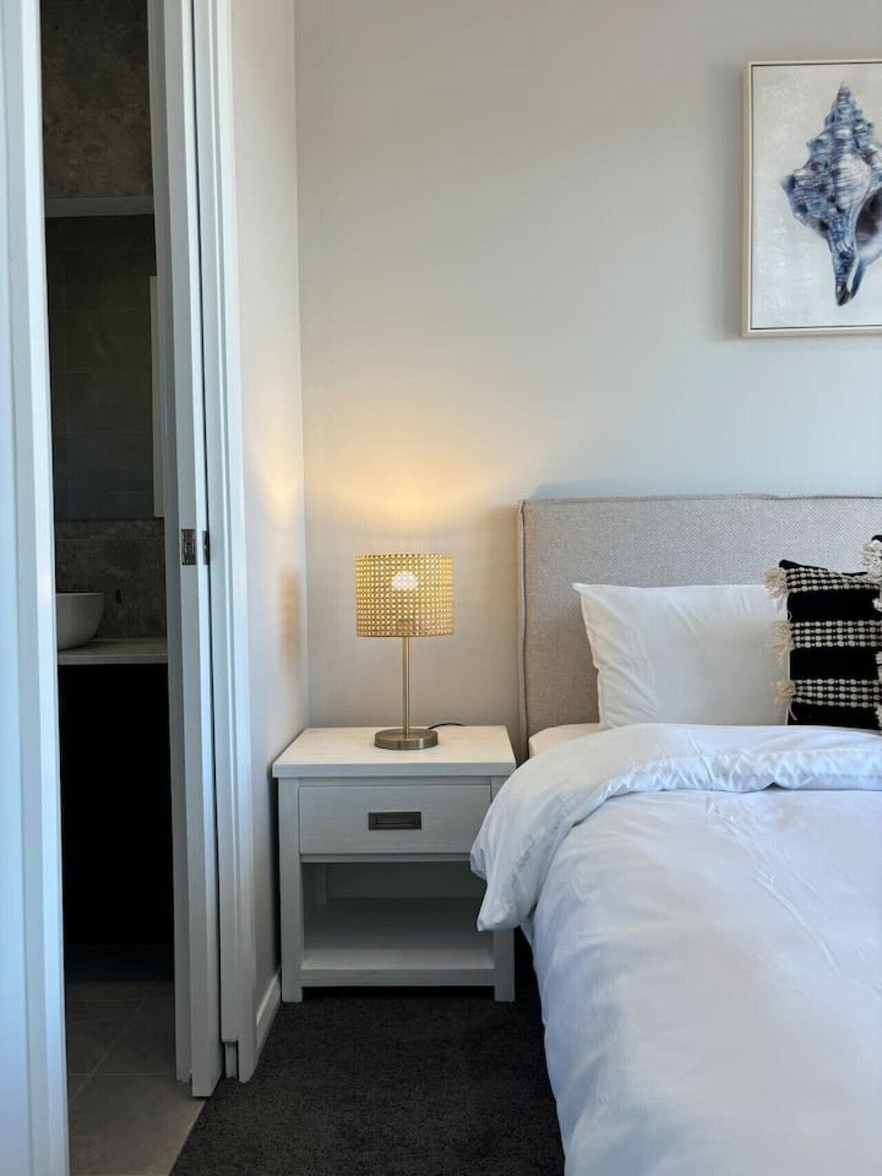 Ein Hotelzimmer mit einem Bett, einem Nachttisch mit Lampe, einem durchgegangenen Tür sichtbar erkennbaren Badezimmer und einem gerahmten Kunstwerk an der Wand.