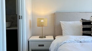 Une chambre d’hôtel avec un lit, une table de chevet avec une lampe, une salle de bain visible à travers une porte ouverte et une œuvre d’art encadrée au mur.