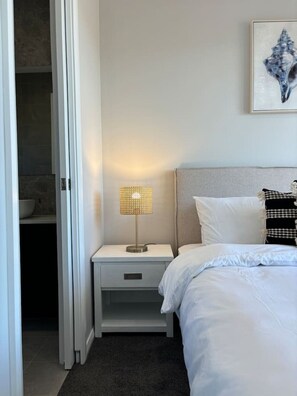 A hotel room with a bed, a nightstand with a lamp, a bathroom visible through an open door, and a framed artwork on the wall.