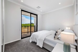 A bedroom with a bed, a nightstand, a lamp, and a sliding glass door leading to a balcony with a view of a vineyard.