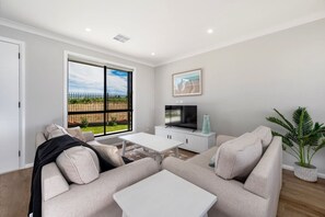 A living room with a sofa, coffee table, TV, and a large window with a view of a fence and greenery.