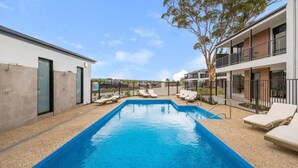 Une piscine avec des chaises longues, un bâtiment doté d’un balcon et un arbre.