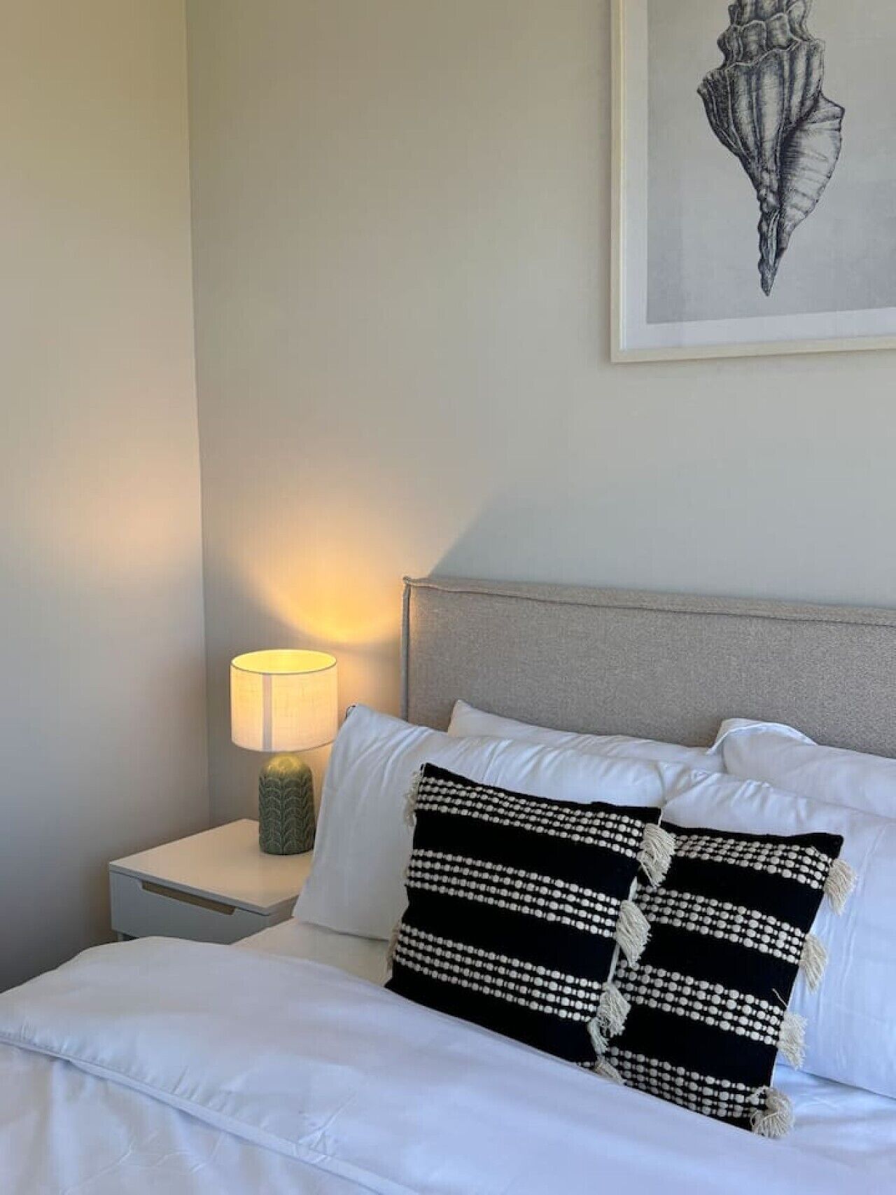 A neatly made bed with white linens and decorative pillows, a bedside lamp, and a framed artwork on the wall.