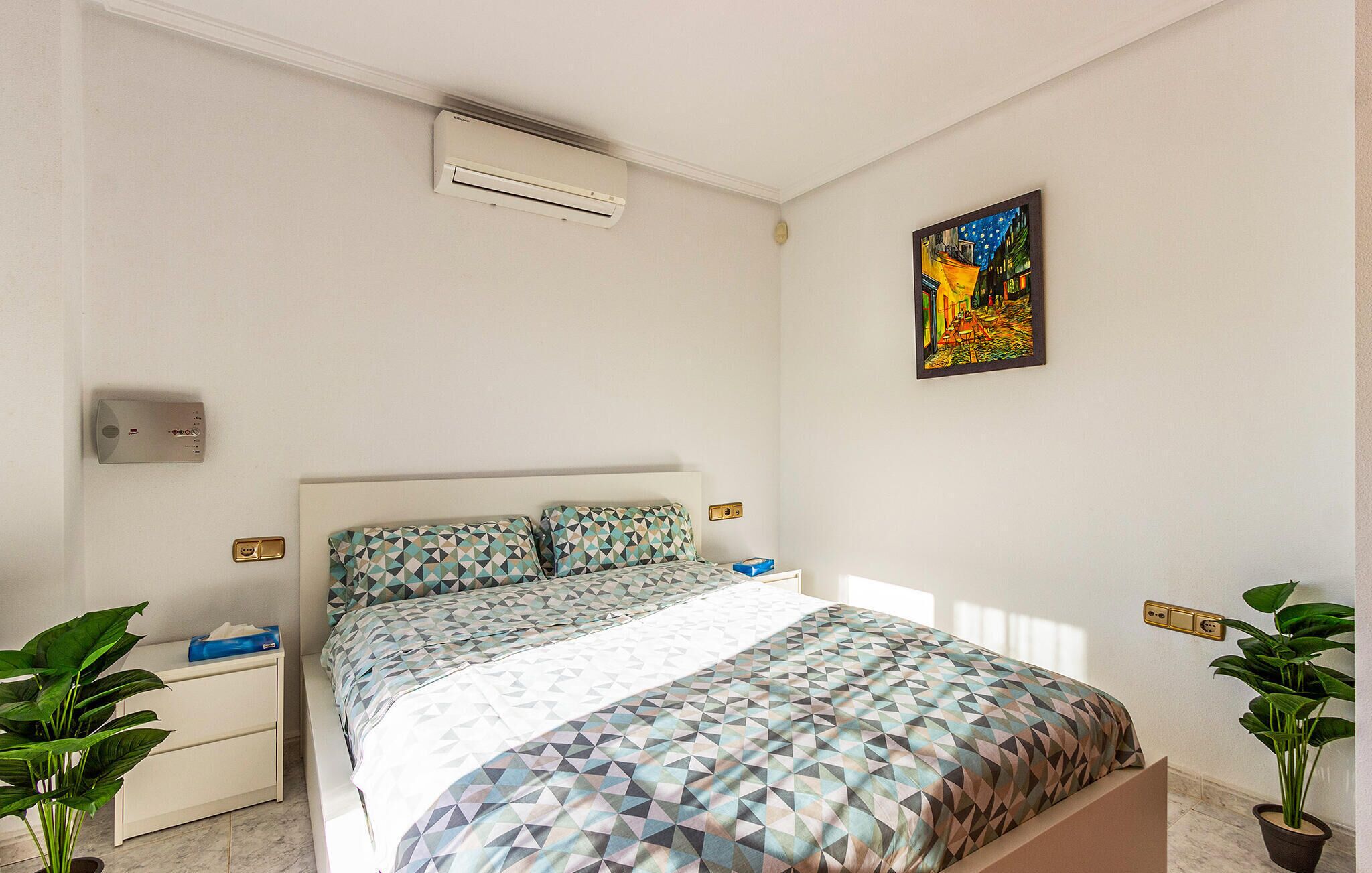 A bedroom with a bed, bedside tables, an air conditioning unit, a painting, and two potted plants.