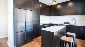 Une cuisine moderne avec des meubles de couleur sombre, un réfrigérateur en acier inoxydable, un îlot central doté de tabourets et un luminaire suspendu.