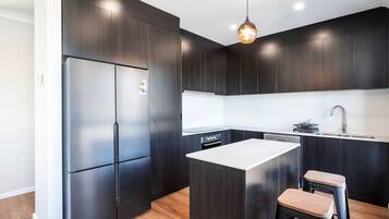A modern kitchen with dark cabinetry, a stainless steel refrigerator, a central island with stools, and a pendant light.