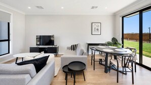 Un salon moderne doté d’un téléviseur à écran plat, de canapés gris, d’une table à manger avec des chaises et d’une grande fenêtre donnant sur l’extérieur.