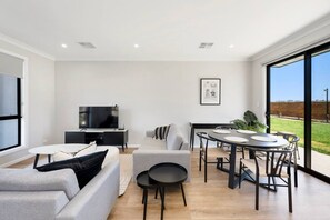 Una sala de estar moderna con televisor de pantalla plana, sofás grises, una mesa de comedor con sillas y un ventanal con vistas al exterior.