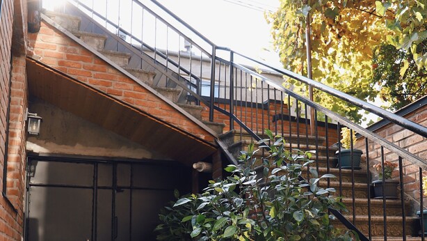 Un edificio de ladrillo con una escalera que lleva a un segundo piso. Hay una planta en maceta en primer plano y una puerta con manija metálica.