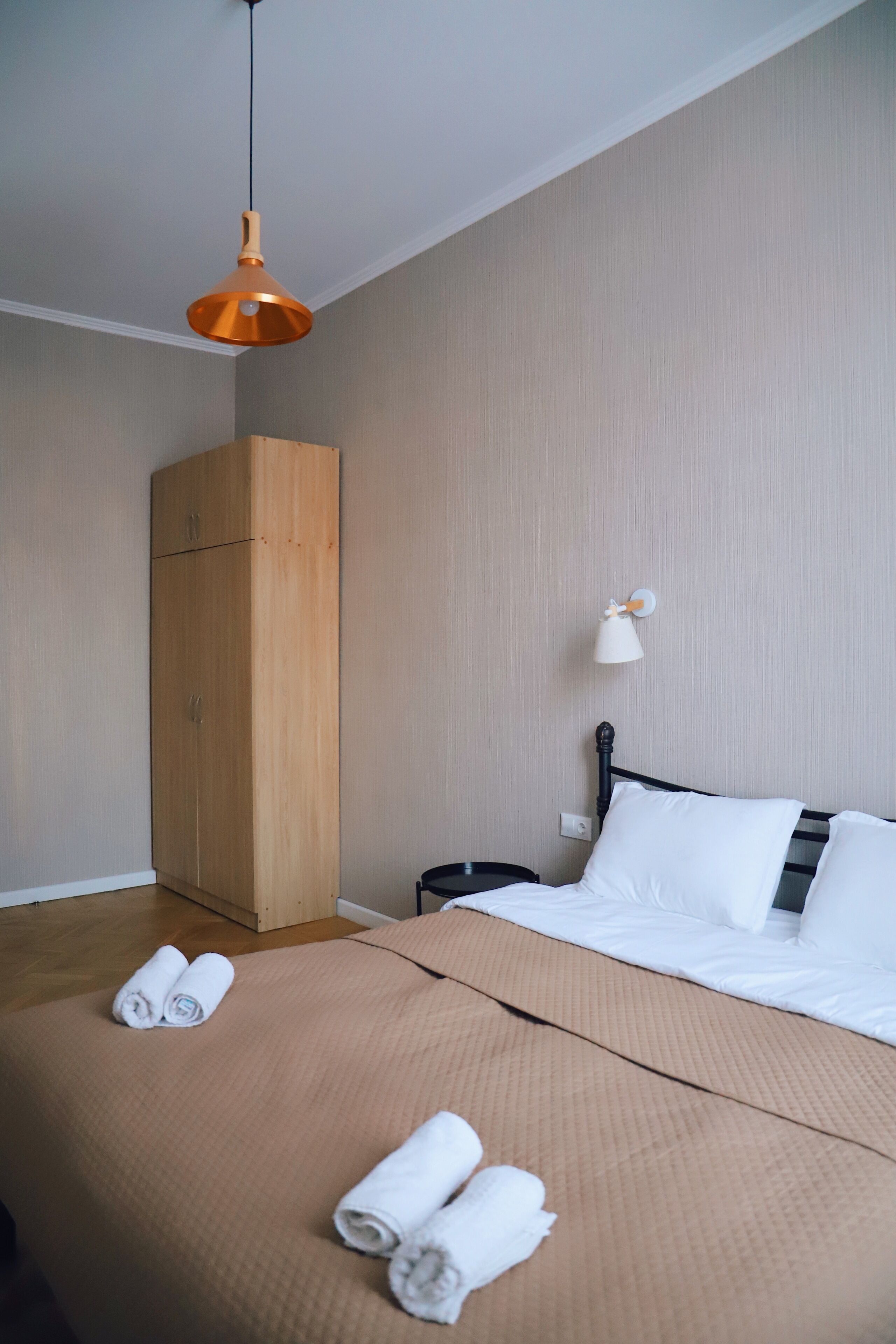 Habitación de hotel con cama, armario de madera, mesita de noche, lámpara y toallas enrolladas sobre la cama.
