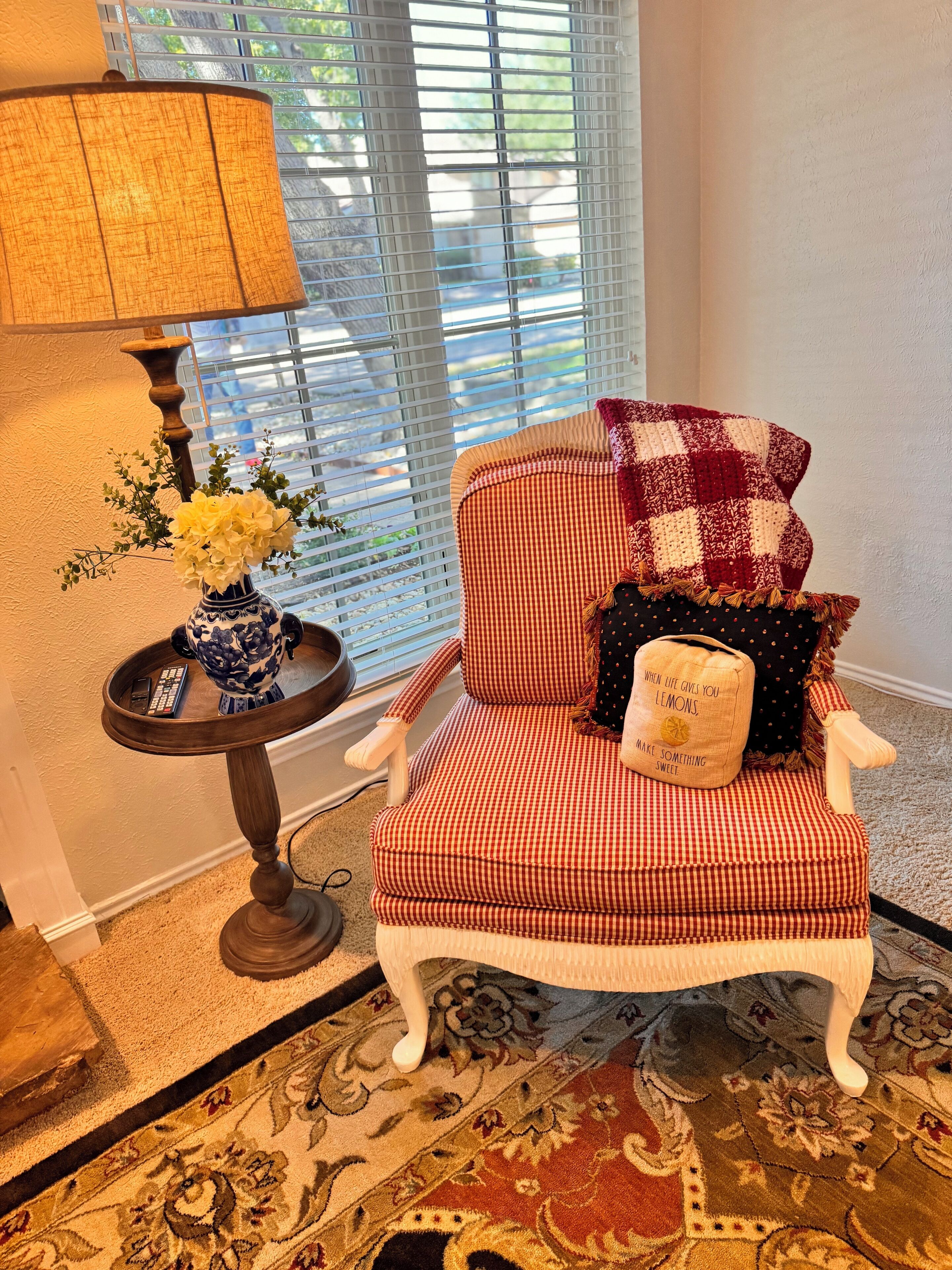 Una habitación acogedora con un sillón a cuadros, una mesita de noche con una lámpara y un jarrón con flores.