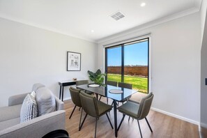 Un espace repas moderne avec une table et des chaises en verre, un canapé et une grande fenêtre donnant sur un champ.
