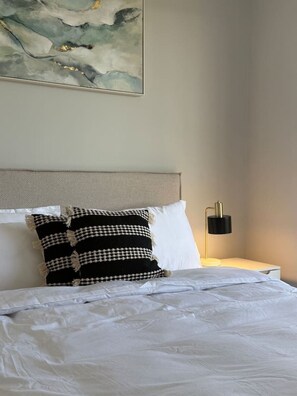 Ein ordentlich bezogenes Bett mit weißen Leinen und zwei Kissen, eines schlicht weiß, das andere mit schwarz-weißer Musterung. Eine moderne Lampe steht auf einem Nachttisch auf der rechten Seite.