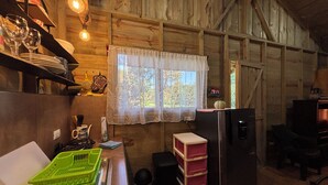 A rustic kitchen with wooden walls, a window with lace curtains, a refrigerator, and a sink with a green dish rack.
