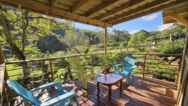 A wooden deck with two blue chairs and a small table, surrounded by lush greenery and trees.