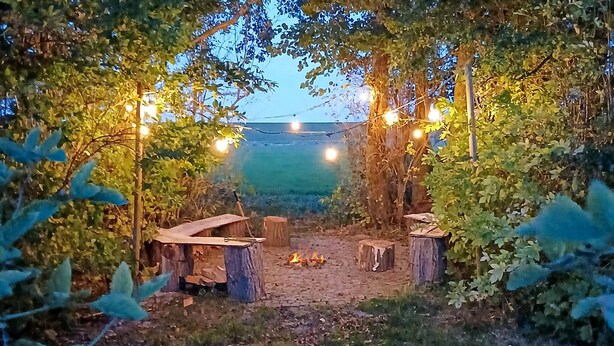 Un área de estar al aire libre con una chimenea, bancos de madera y luces colgantes.