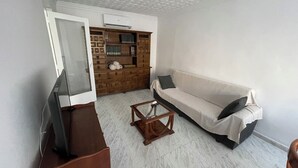 A living room with a sofa, a coffee table, a TV stand, and a wooden cabinet with books and rolled-up towels.