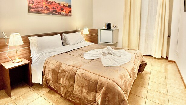 A hotel room with a bed, a nightstand, a lamp, a painting on the wall, and a window with curtains. - Vitti's home Verona 5 (Verona)
