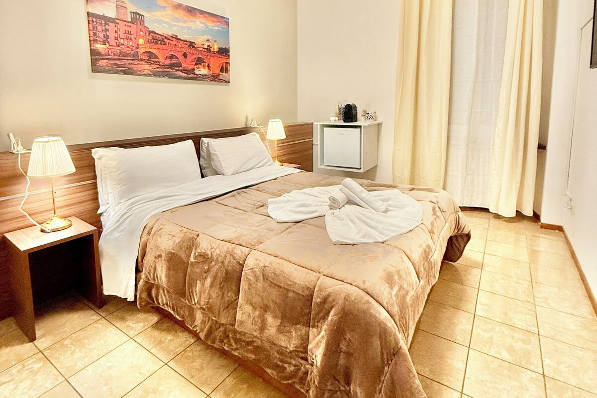 A hotel room with a bed, a nightstand, a lamp, a painting on the wall, and a window with curtains.
