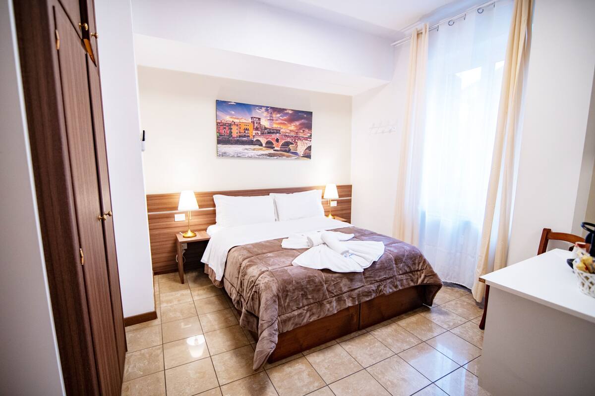 A hotel room with a bed, a nightstand, a desk, and a painting on the wall.
