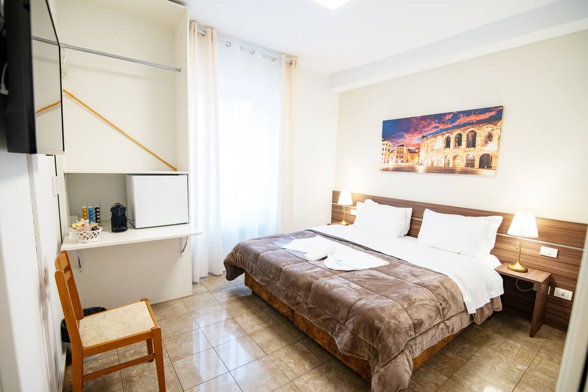A hotel room with a bed, a chair, a desk, a painting, and a nightstand.